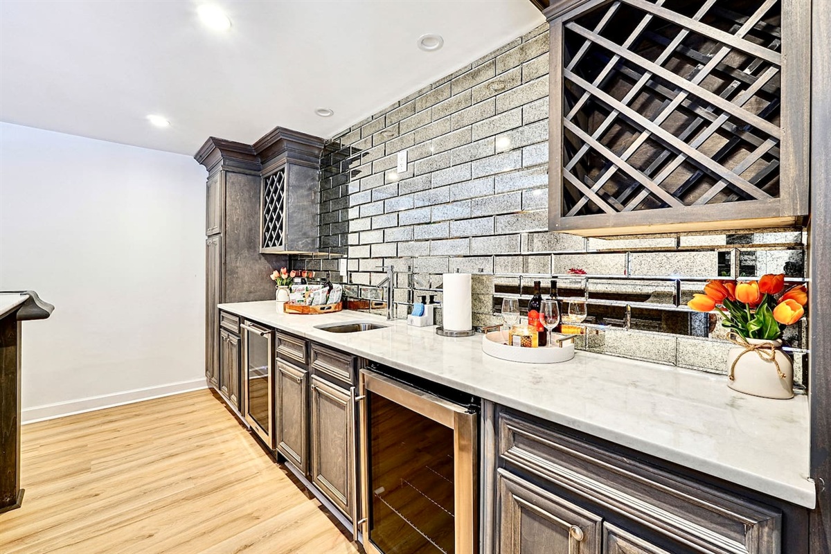 Basement kitchenette located to the left at the bottom of the stairs, featuring a sleek bar with seating, sink, two coolers, wine cabinetry, and modern gray cabinets with a matching backsplash, convenient as it is right next to the game room!
