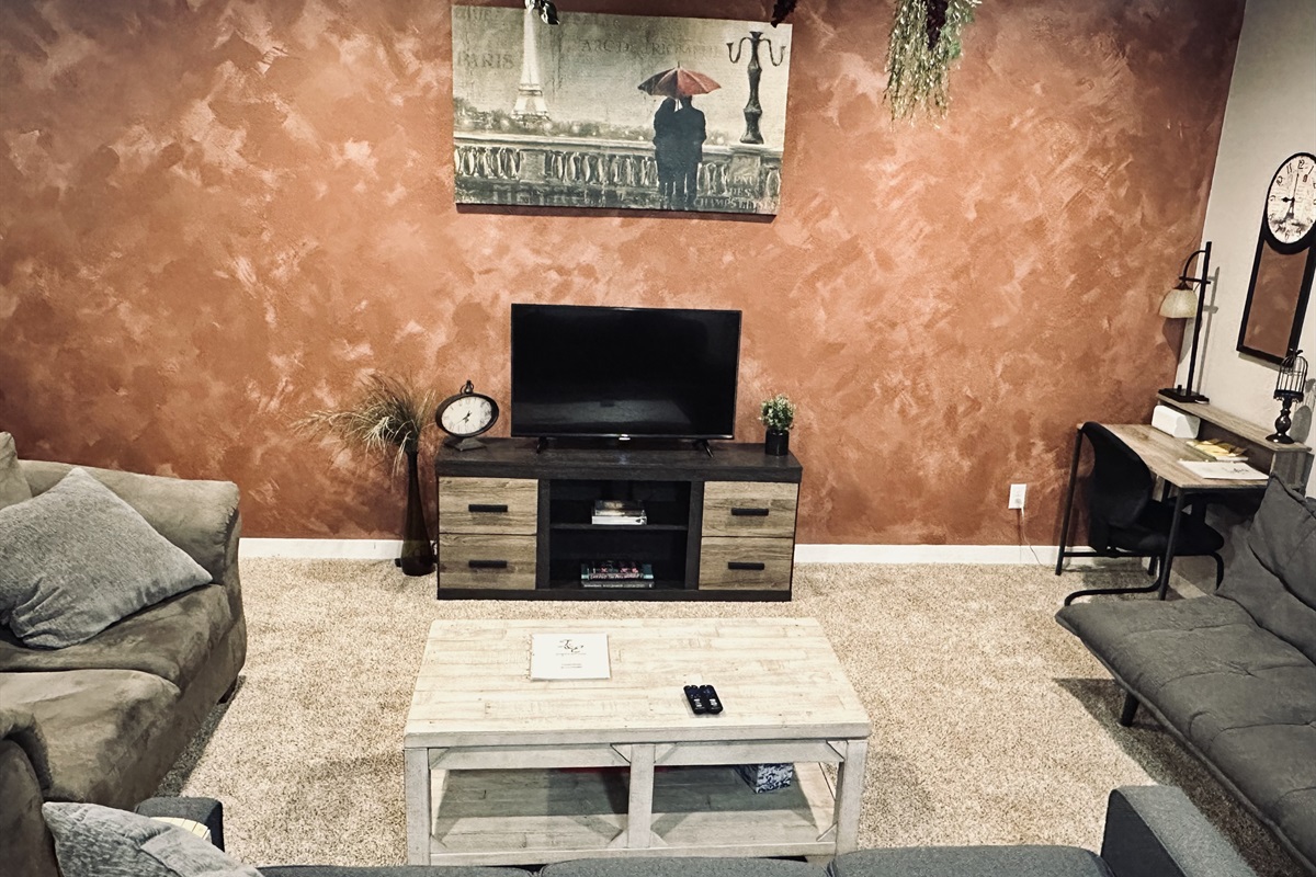 A cozy living room featuring a burnt orange textured wall, a central wooden coffee table, a television on a dark wood media console with decorative elements, a comfortable gray sofa, and a workspace with a desk and chair in the corner.