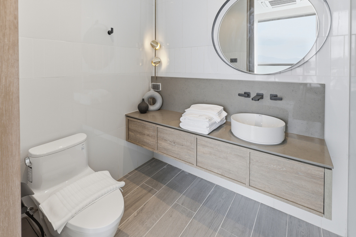 Bathroom with wooden vanity, round mirror, white vessel sink, toilet, and ocean view through window.