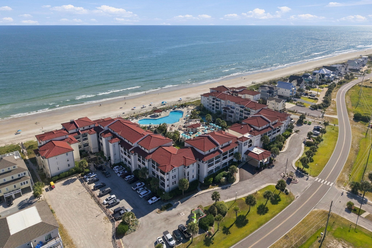 Aerial view of the resort grounds featuring terracotta-roofed residential wings, central pool amenities, parking areas, and adjacent beach frontage bordering coastal access roads.   
