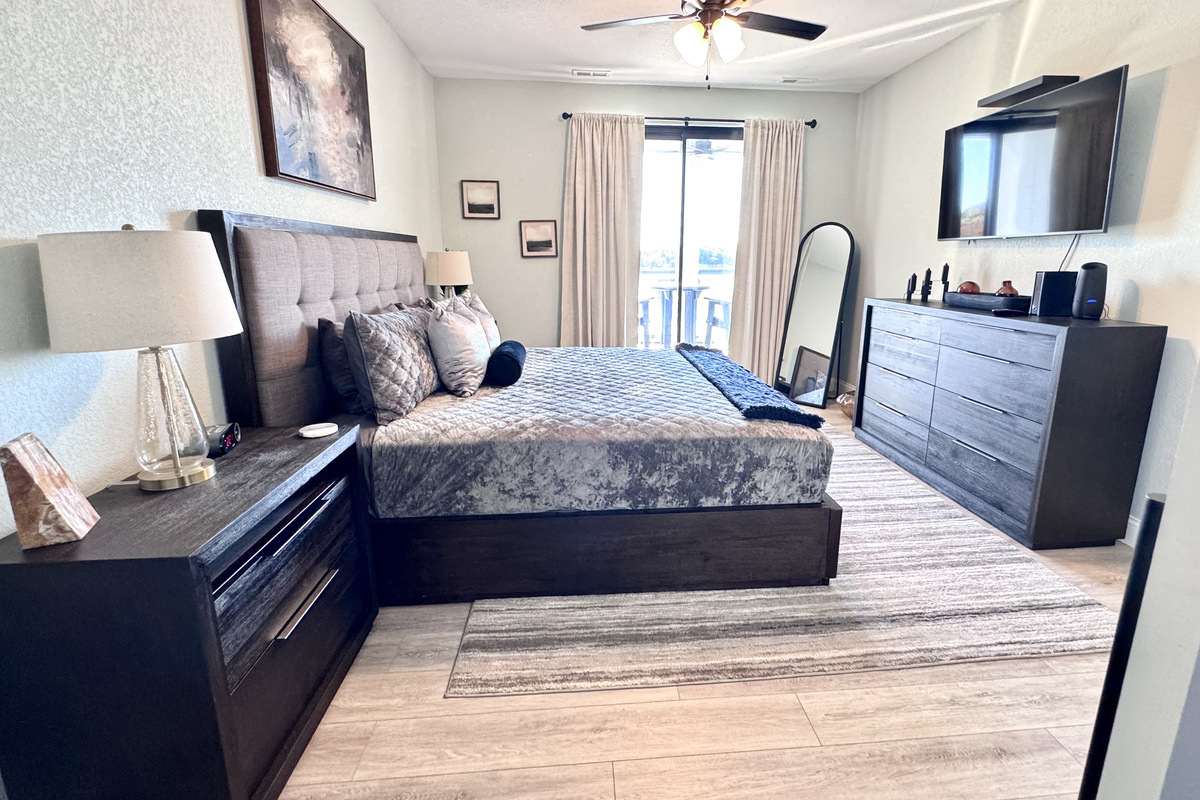 Primary Bedroom with a lakeside view and balcony access. Includes a Smart TV, large dresser, full-length mirror, two nightstands, table lamps, all bed linens, extra pillows, blanket, and cell phone/watch charging stations.