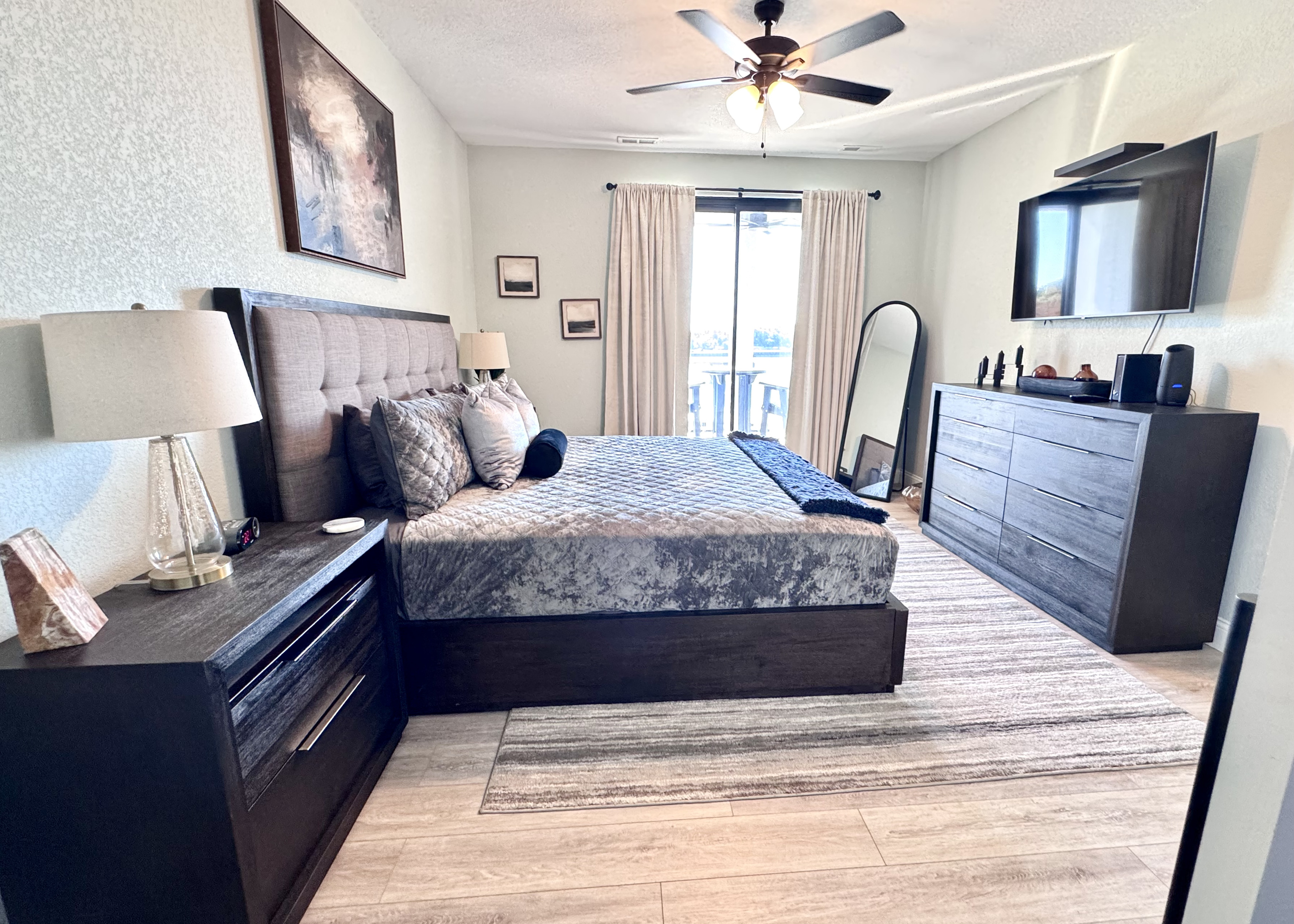 Primary Bedroom with a lakeside view and balcony access. Includes a Smart TV, large dresser, full-length mirror, two nightstands, table lamps, all bed linens, extra pillows, blanket, and cell phone/watch charging stations.