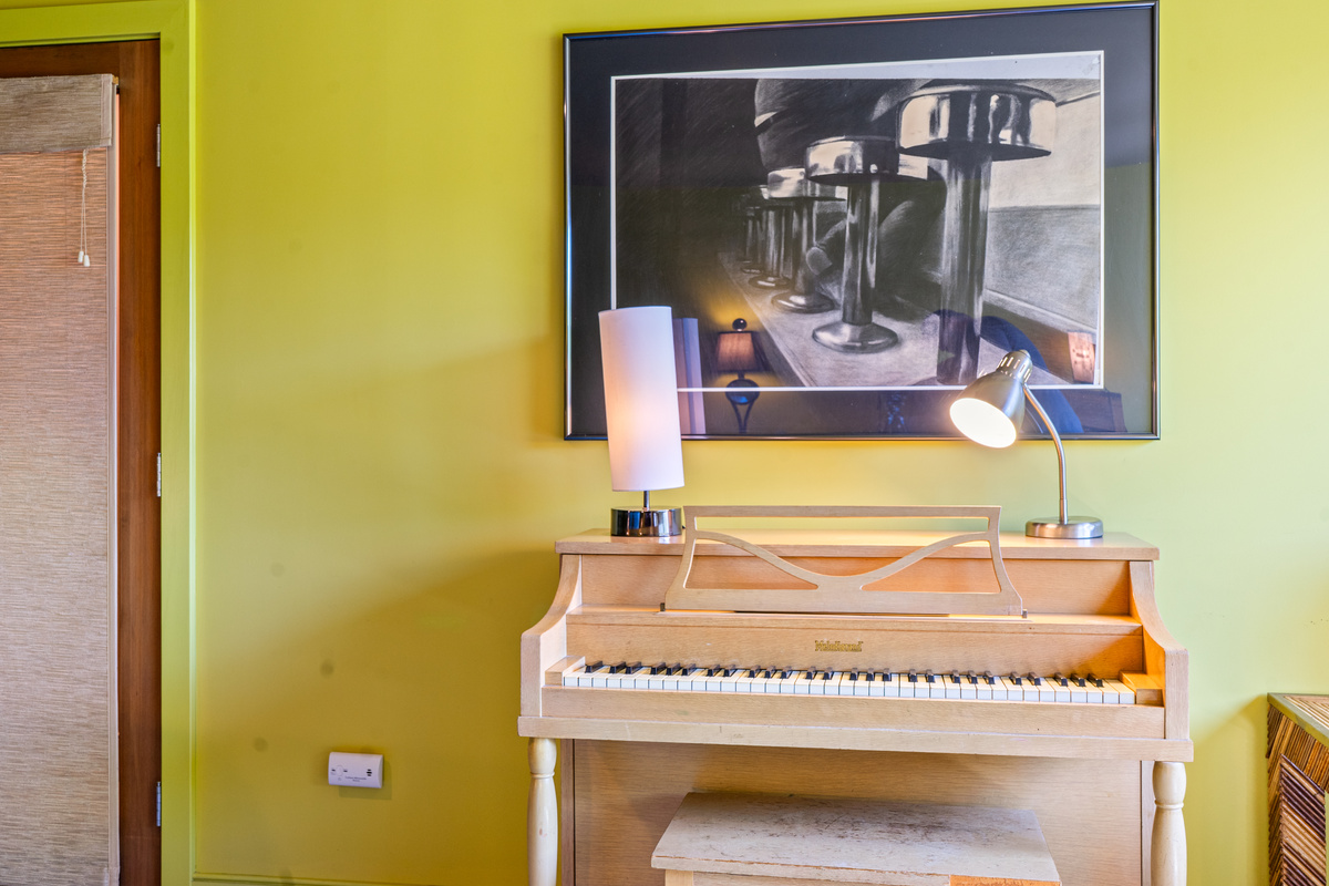 Artistic detail in "The Crag's" Base Camp bedroom. A blonde wood piano, warm directional lamps, and striking framed art create a unique, creative corner inside this ground-floor retreat near Acadia.