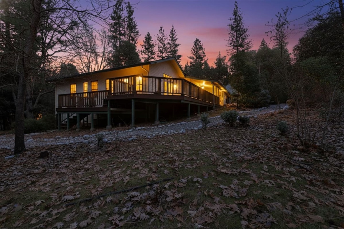Cabin exterior at sunset with a wraparound deck, wooded setting, pine tree surroundings, and warm evening lighting for a peaceful mountain retreat.