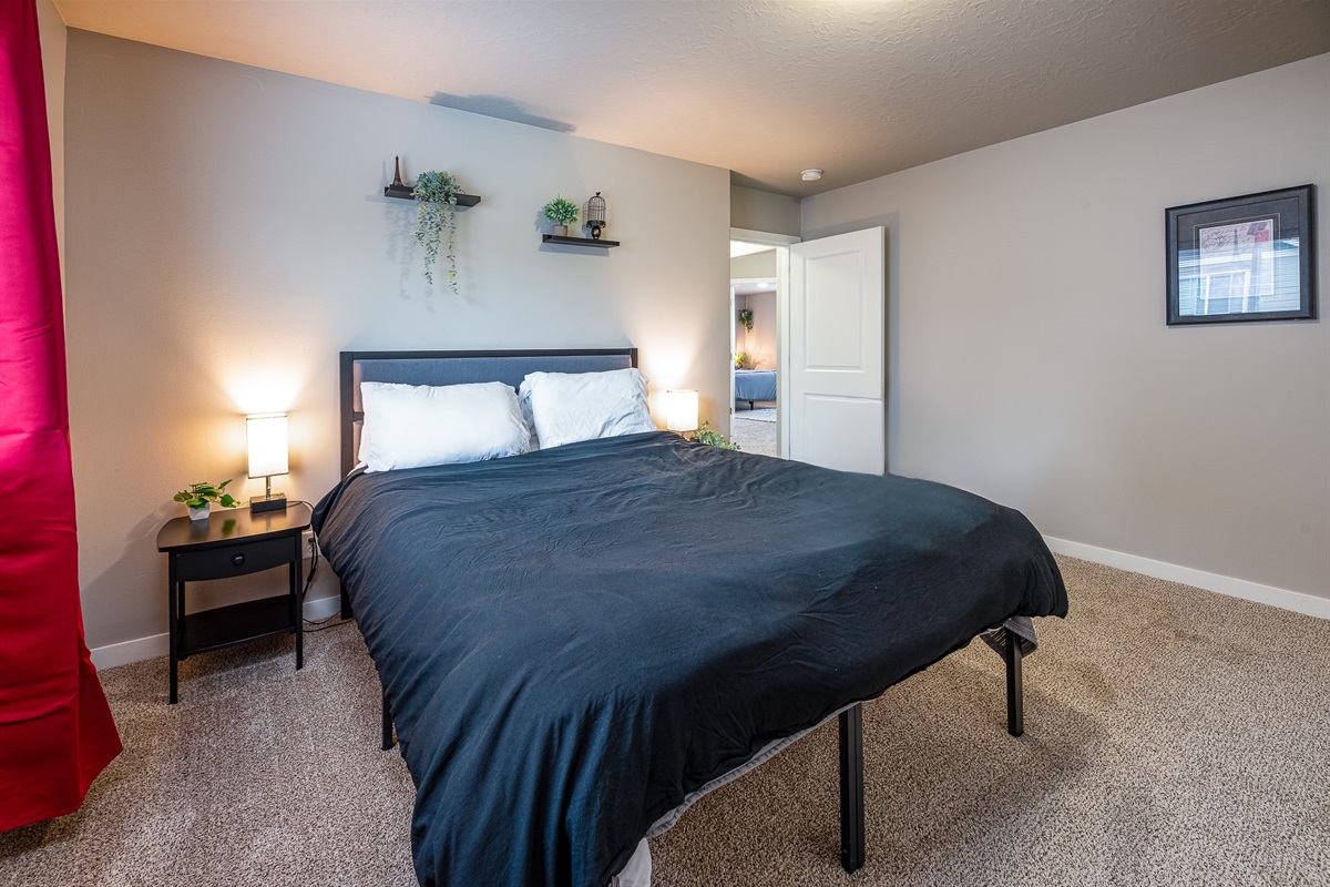 A cozy bedroom featuring a black bedspread over a bed with white pillows, a small black nightstand with a lamp and a potted plant beside it, and shelves with decorative plants mounted on the wall, with a doorway leading to another room visible in the background.