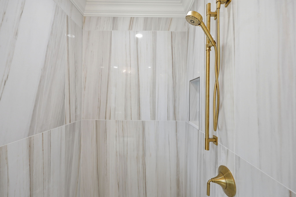 Glass-enclosed walk-in shower with marble-style tile walls and gold rainfall shower fixtures in a bright modern bathroom