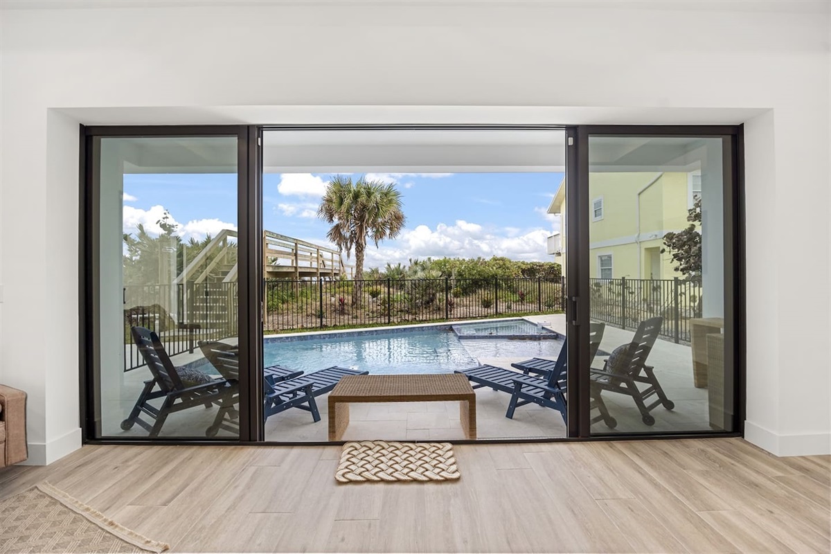 The event room opens to a stunning view with a table and chairs, a glass sliding door revealing a sparkling swimming pool and lush greenery outside, offering a seamless blend of indoor comfort and outdoor beauty.