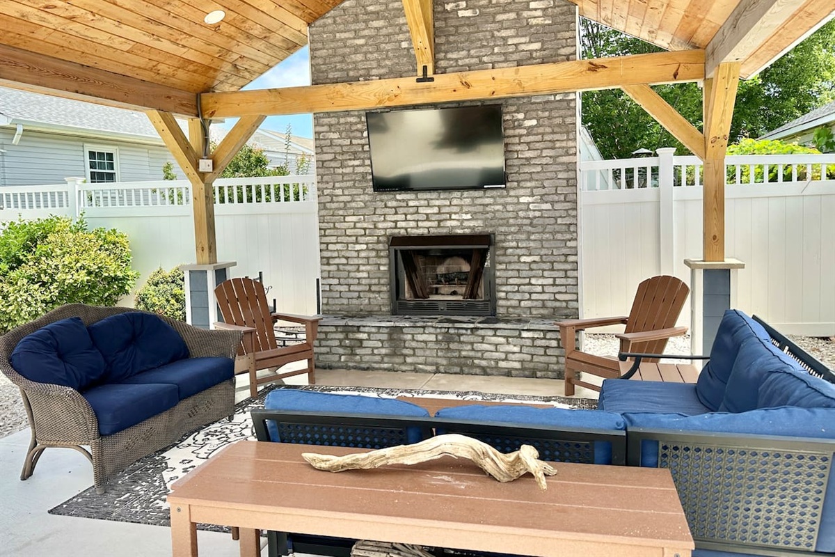 This shaded pavilion features a brick fireplace and a large flat-screen TV for outdoor entertainment. The seating area includes navy blue sofas and wooden Adirondack chairs under a vaulted ceiling.