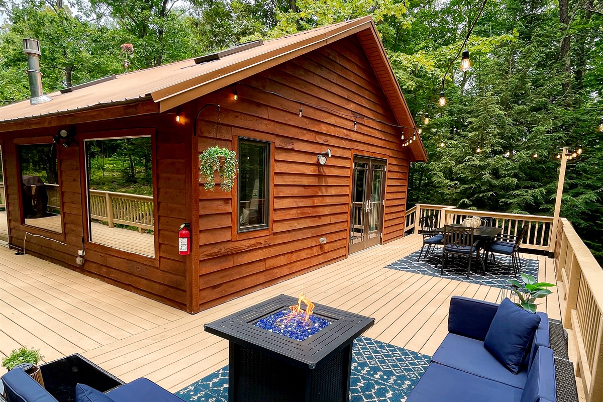 Main cottage back deck feature lounge couch with propane fire pit, 6 person dining table lit by string lights. Wooded views. 