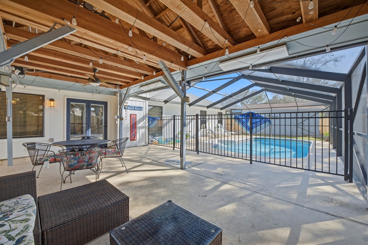 Designed for effortless indoor-outdoor living, this expansive covered patio features lounge seating, a dining area, and overhead lighting beneath stunning wood beams. The screened structure opens to a private pool area, ideal for relaxing or hosting.