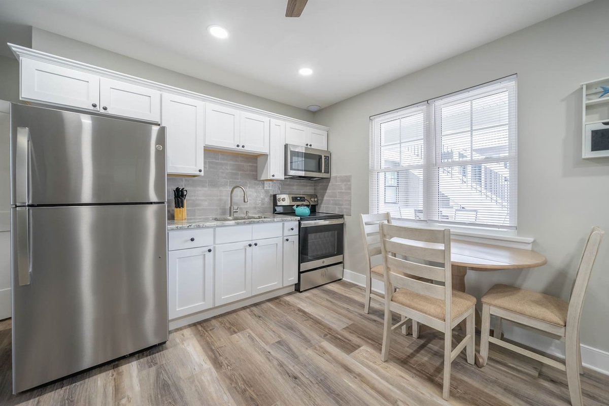 Bright, modern kitchen with stainless steel appliances, white cabinetry, and ample counter space. A small dining table by the window creates a comfortable spot for meals, with natural light enhancing the clean, open feel of the space.