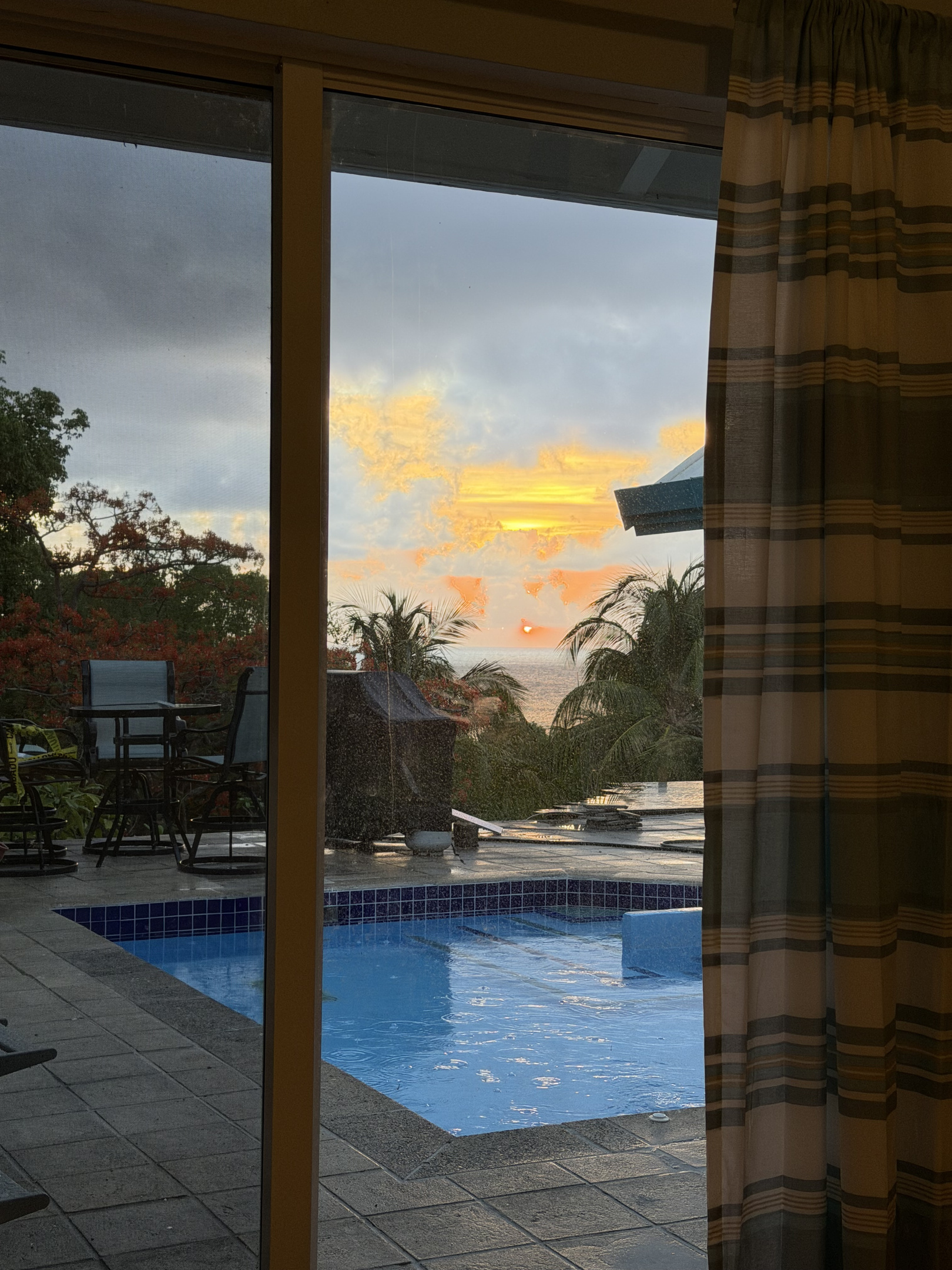 The primary bedroom sliding glass door opens onto a spacious tiled patio featuring a shimmering, bright blue swimming pool. Beyond the pool area is lush tropical greenery and a peaceful glimpse of the open sea.