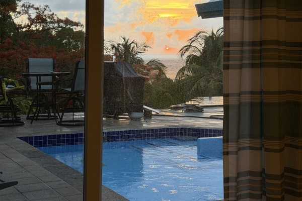 The primary bedroom sliding glass door opens onto a spacious tiled patio featuring a shimmering, bright blue swimming pool. Beyond the pool area is lush tropical greenery and a peaceful glimpse of the open sea.