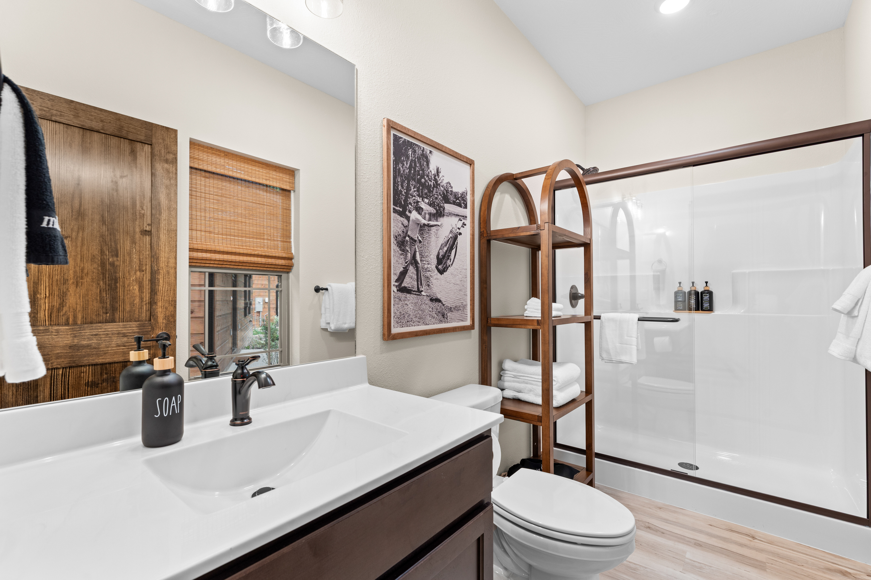 The main level bedroom ensuite has a glass walk-in shower, a ladder of fresh towels, and a wink of vintage golf on the wall.