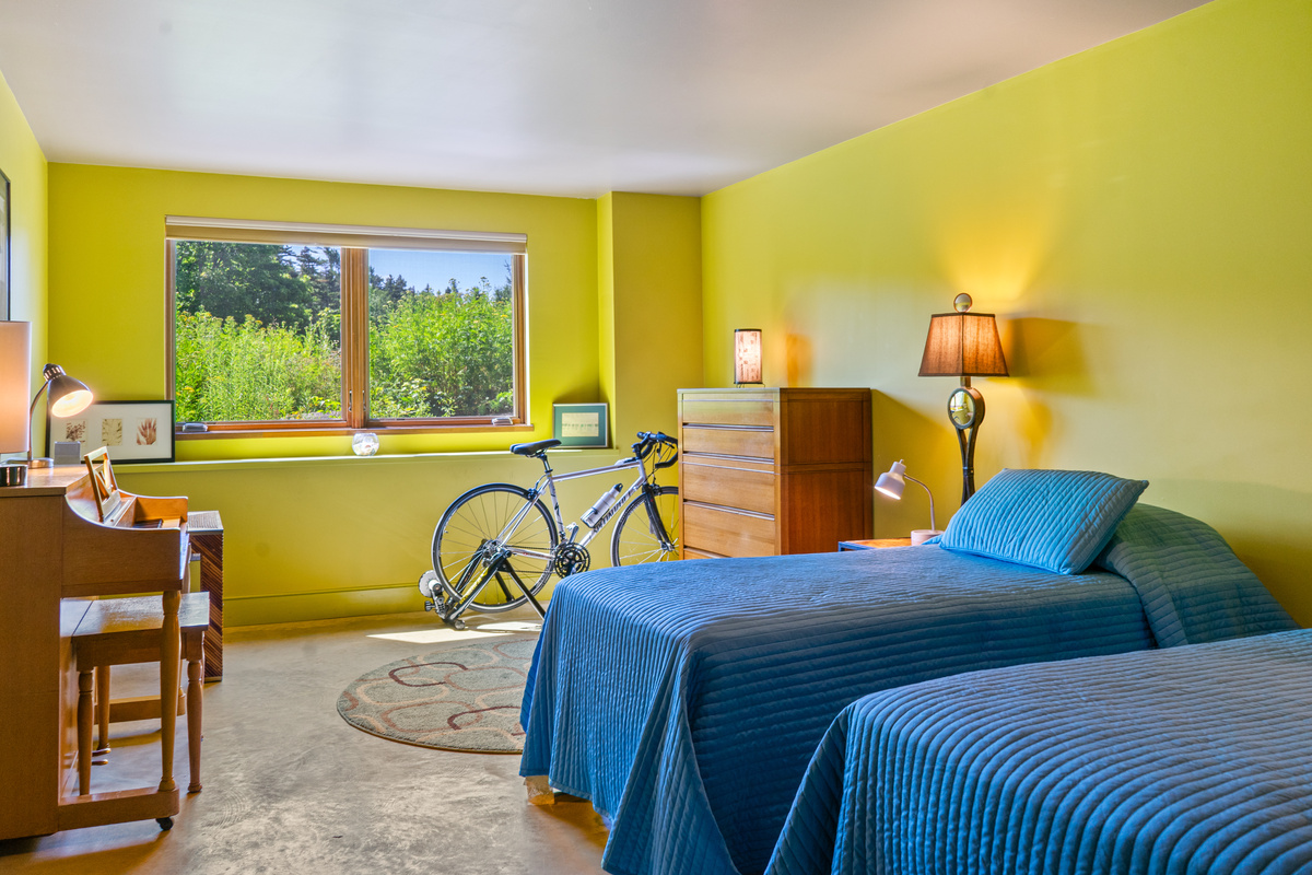 "The Crag's" Base Camp bedroom features twin beds, dedicated storage, a piano, and a large window looking onto private, custom-landscaped front grounds. A vibrant ground-floor retreat ideal for kids, friends, or adventurers exploring Acadia.