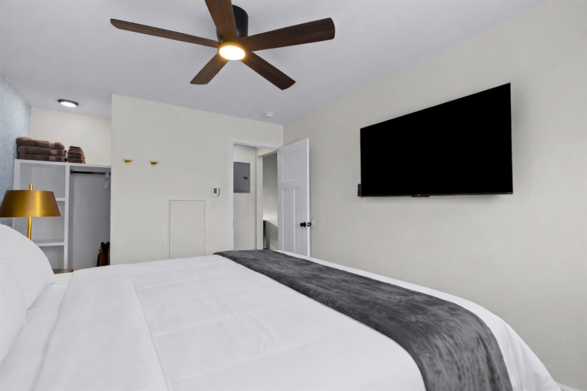 Kick back in this cozy bedroom with a wall-mounted smart TV, ceiling fan, and plush bedding—perfect for winding down and enjoying a quiet night in.