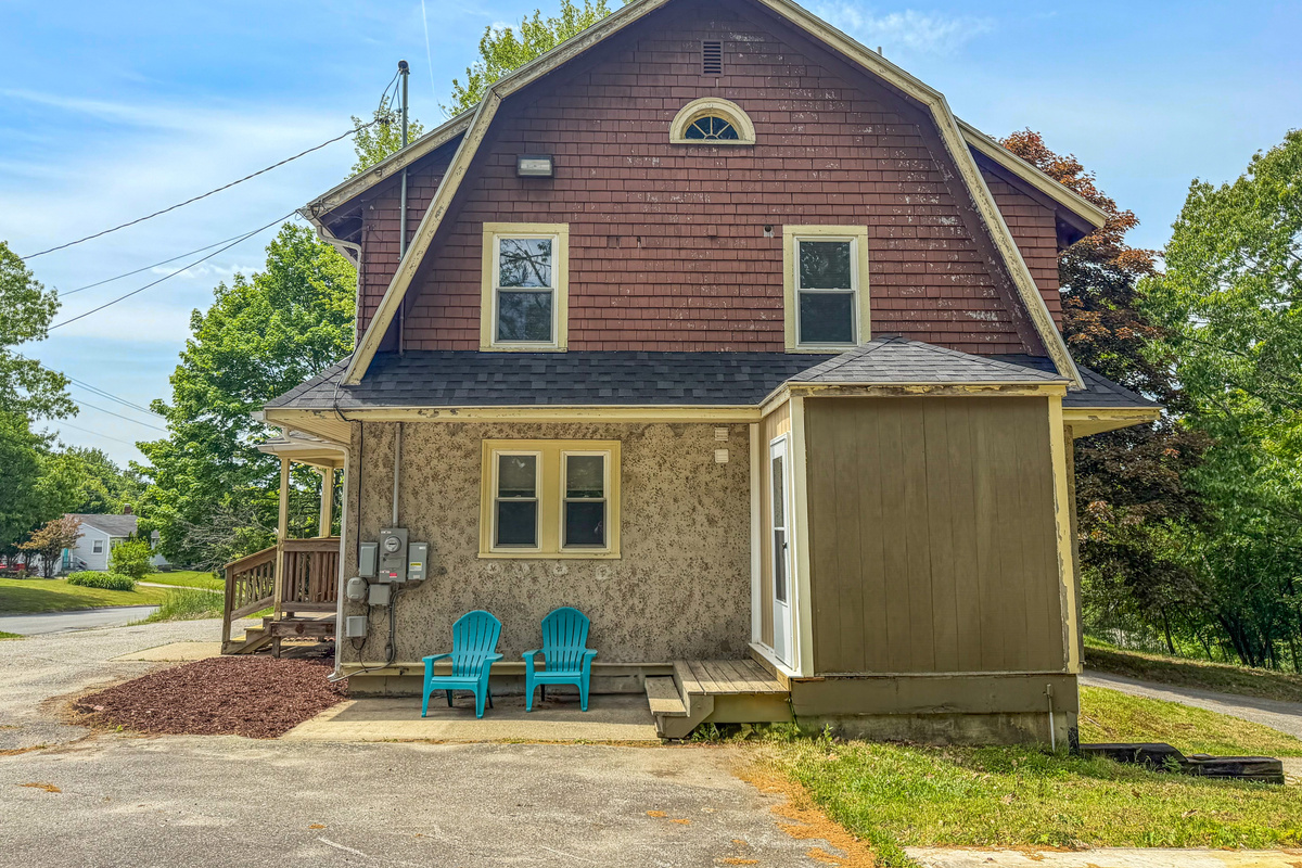 The side exterior profile showcases the main mudroom entry door and wooden steps. Two turquoise Adirondack chairs sit directly on the concrete pad off the driveway, offering a convenient casual outdoor seating area right at your primary entrance.