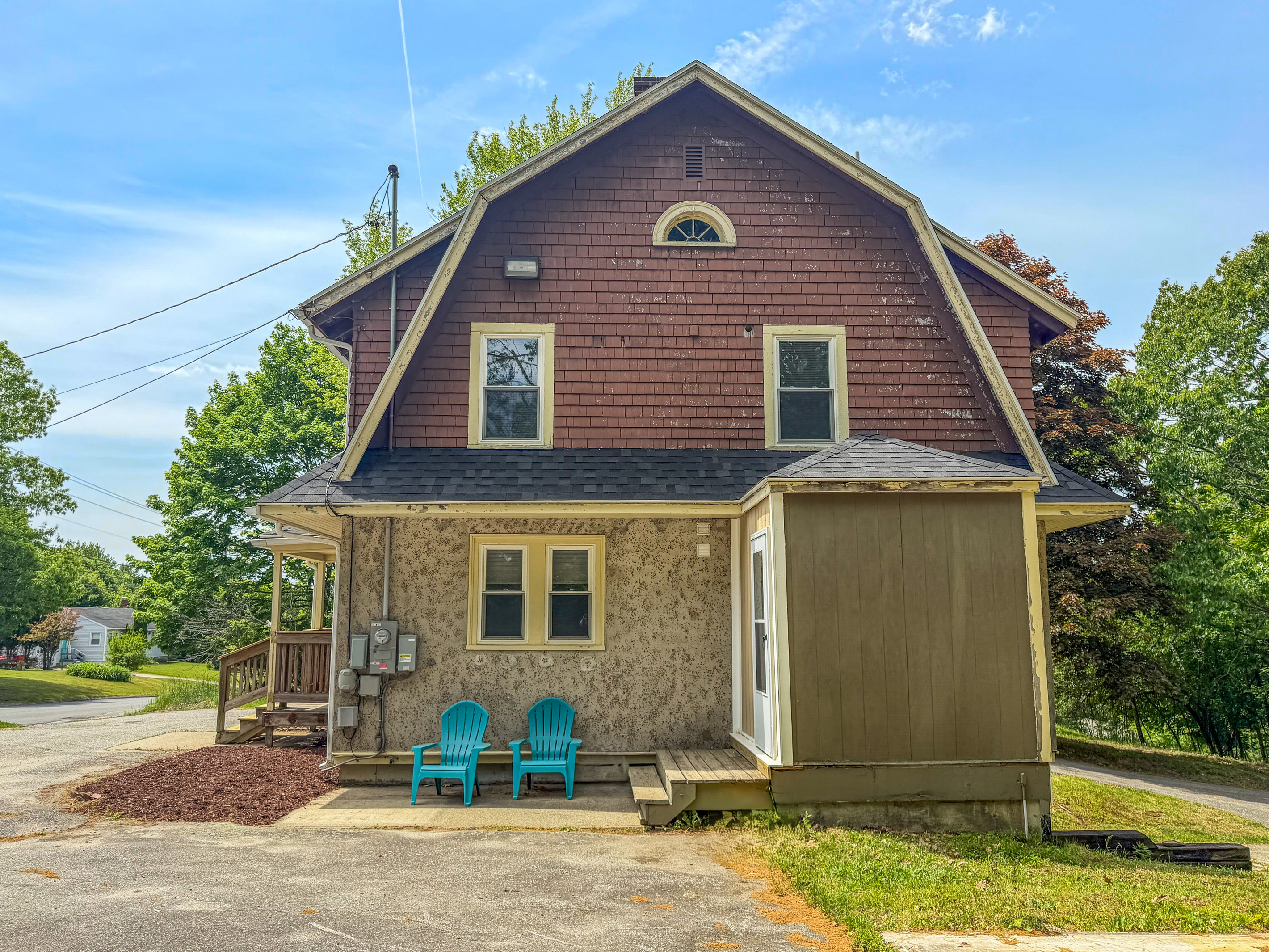 The side exterior profile showcases the main mudroom entry door and wooden steps. Two turquoise Adirondack chairs sit directly on the concrete pad off the driveway, offering a convenient casual outdoor seating area right at your primary entrance.