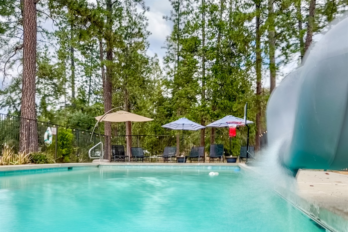 A fenced pool area features a water slide cascading into the shimmering water, surrounded by lounging chairs and colorful umbrellas providing shade and relaxation.