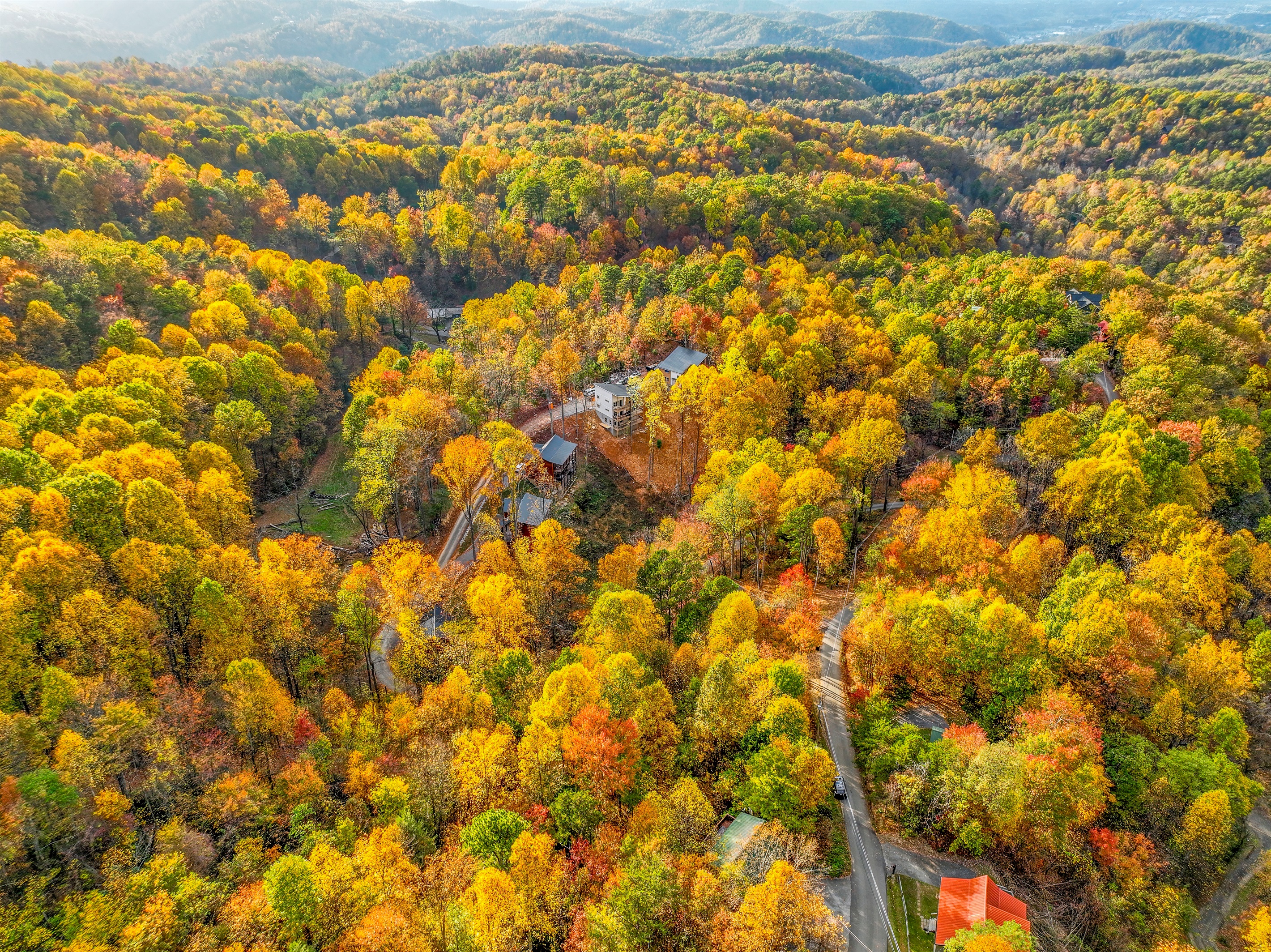 An aerial view of a serene autumn landscape, where vibrant hues of gold, orange, and red blanket the forest. Cozy cabins nestle among the trees, with majestic mountains rising in the distance.