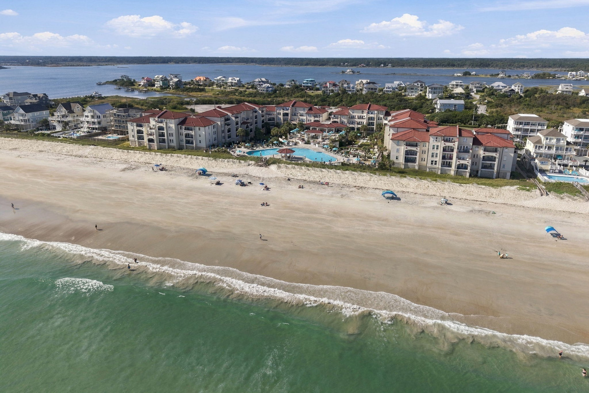 Elevated beachfront view showing the expansive shore, ocean surf, pool area, and resort buildings framed by the sound.