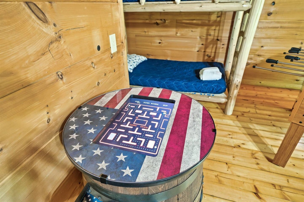 This cozy game corner showcases a barrel-style arcade table decorated with an American flag theme, accompanied by a nearby set of bunk beds, blending fun with cabin-style comfort.