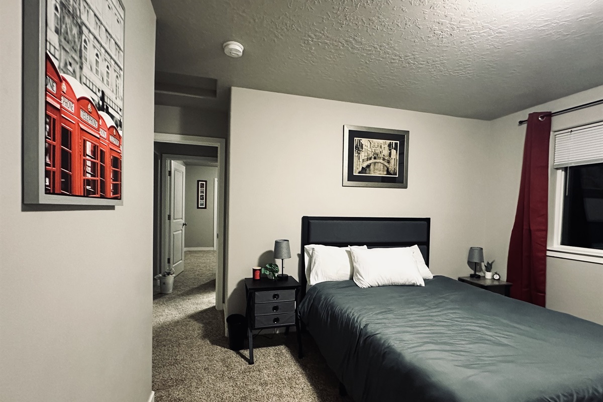 A cozy bedroom features a neatly made bed with gray bedding and white pillows, a bedside table with a small lamp and decorative plant, two wall art pieces including a photo of red telephone booths and a black-and-white city scene, and a doorway leading to a brightly lit hallway.