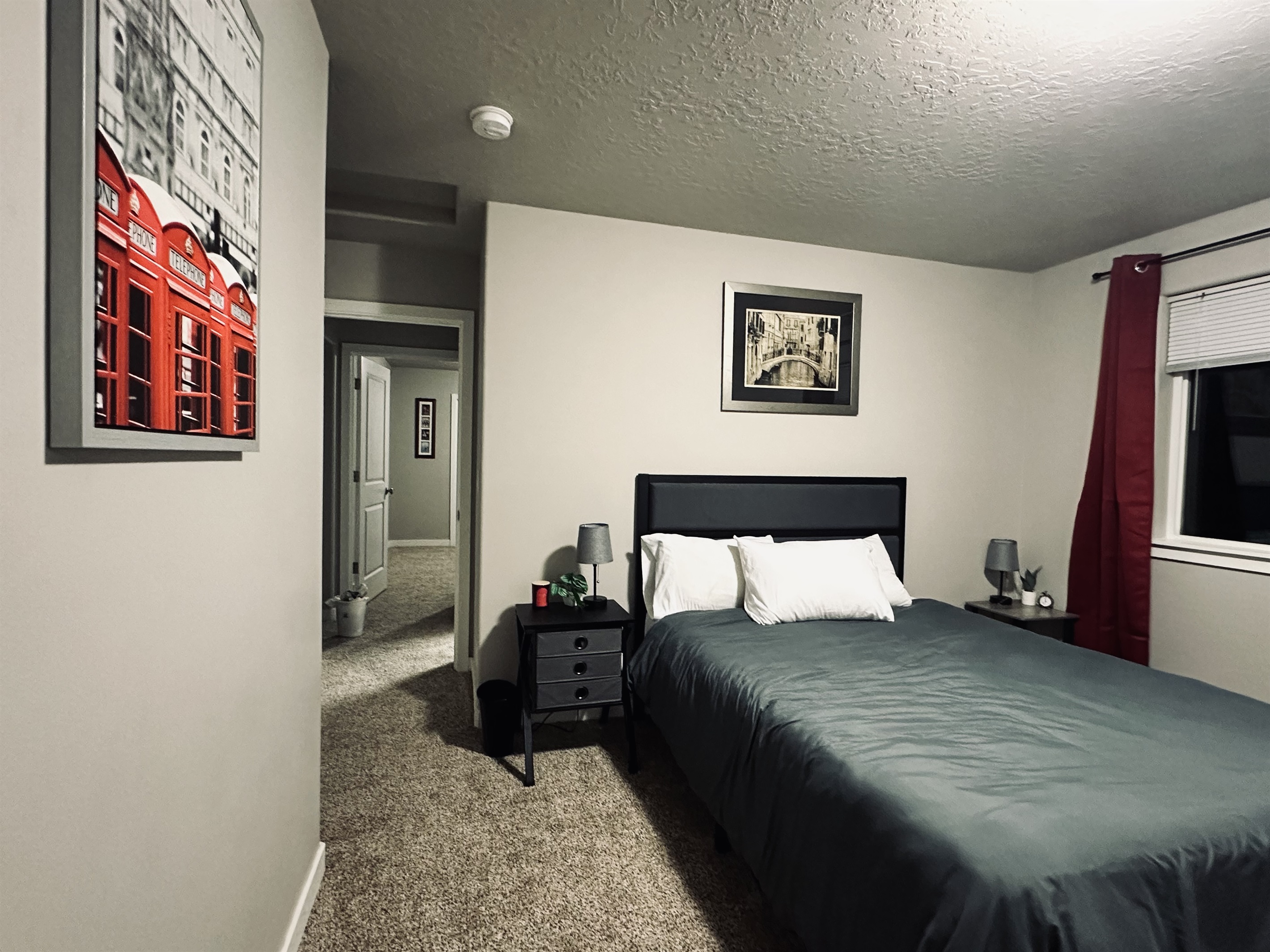 A stylish bedroom features a gray bedspread on a bed with white pillows, a black nightstand with a lamp, and a wall adorned with a framed print of red phone booths, while a doorway leads to a carpeted hallway.