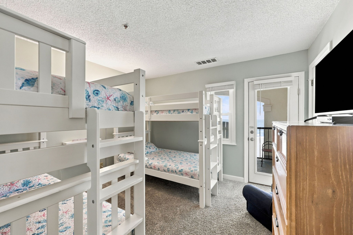 Bright bunk bedroom featuring two twin bunk bed sets with coastal seashell-patterned bedding, a warm wood dresser, wall-mounted TV, and a glass door opening directly to a private ocean-view balcony.   