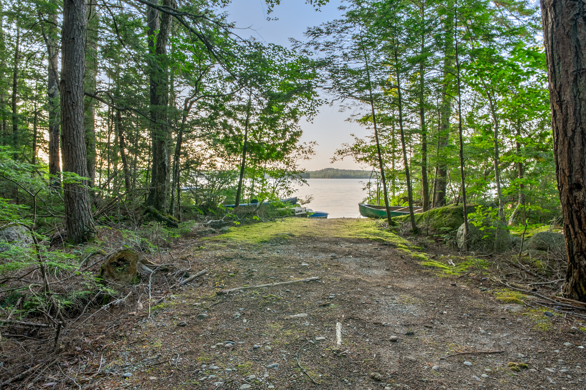 Approaching the lake down the trailer path at Loonrise Point! Packed dirt and stone track leads straight to the water, passing stored kayaks and a canoe. Prime wade-in entry for launch and trailer staging on Beech Hill Pond in Otis, ME.