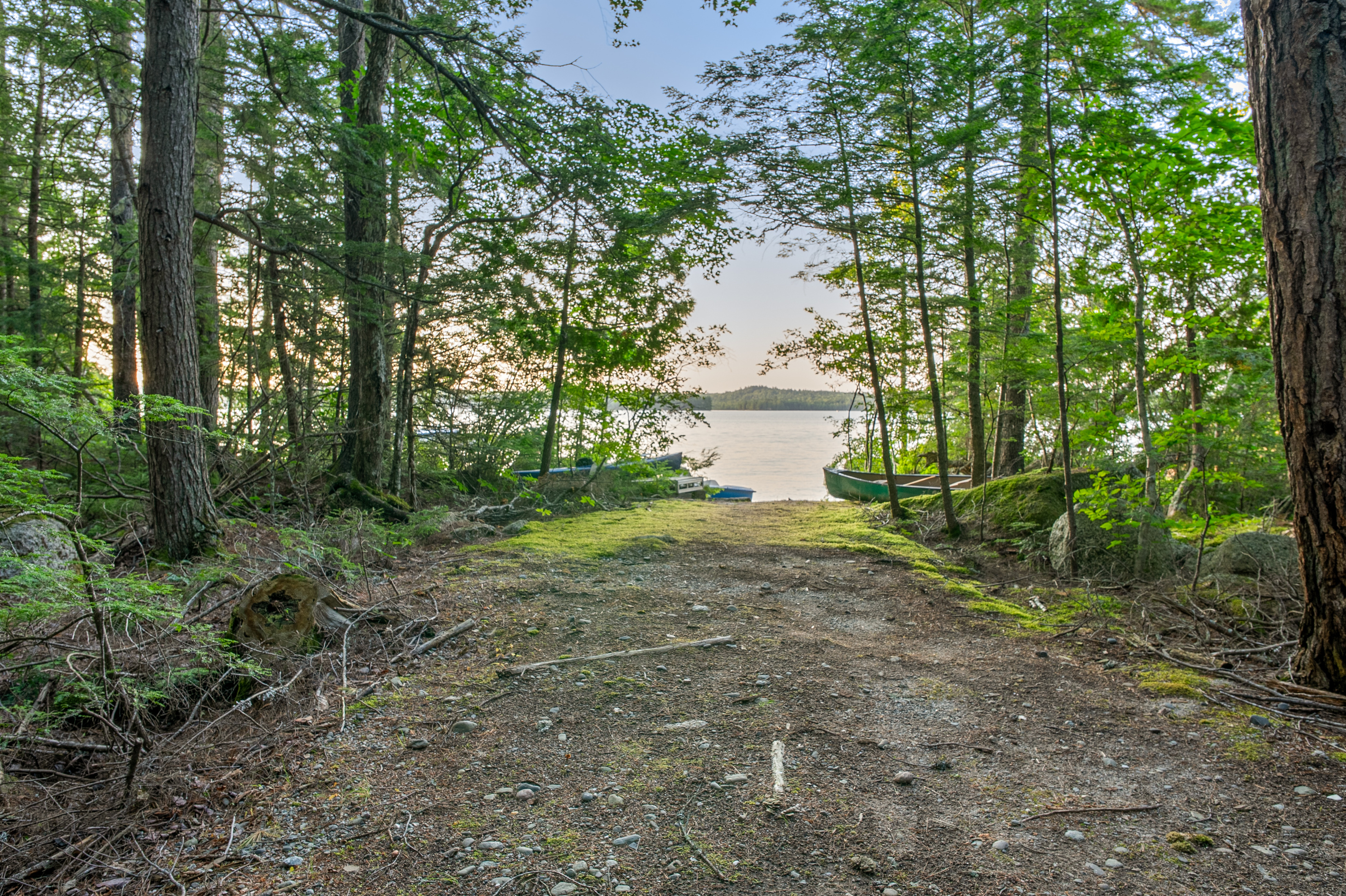 Approaching the lake down the trailer path at Loonrise Point! Packed dirt and stone track leads straight to the water, passing stored kayaks and a canoe. Prime wade-in entry for launch and trailer staging on Beech Hill Pond in Otis, ME.