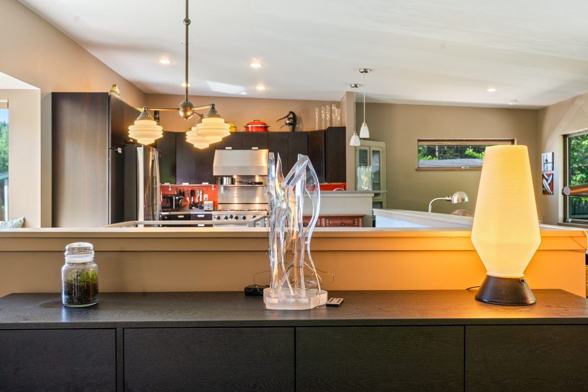 Thoughtful design details at "The Crag." Looking across the dark wood credenza toward the open gourmet kitchen, highlighting warm ambient accent lighting, modern art sculptures, and mid-century fixture details.