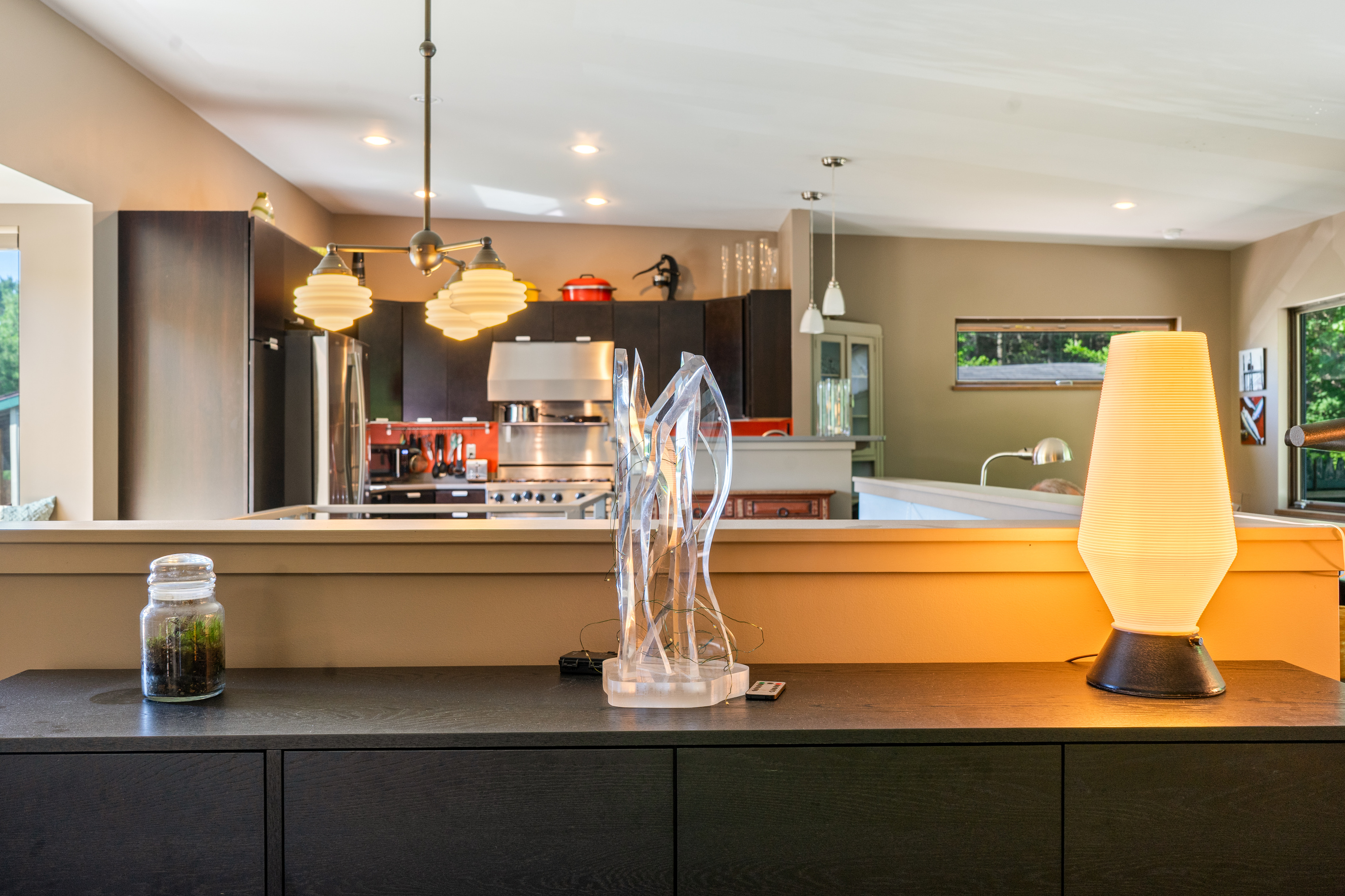 Thoughtful design details at "The Crag." Looking across the dark wood credenza toward the open gourmet kitchen, highlighting warm ambient accent lighting, modern art sculptures, and mid-century fixture details.