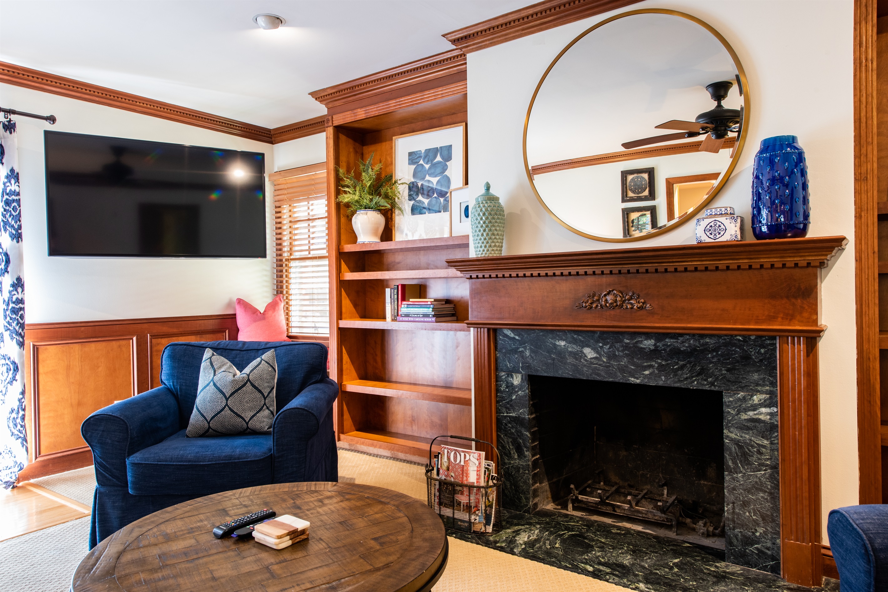 Cozy Airbnb in Louisville, KY, on the Bourbon Trail. Warm fireplace, wooden mantel, built-in shelves, blue armchair, round mirror, and TV. Perfect for a charming getaway! 