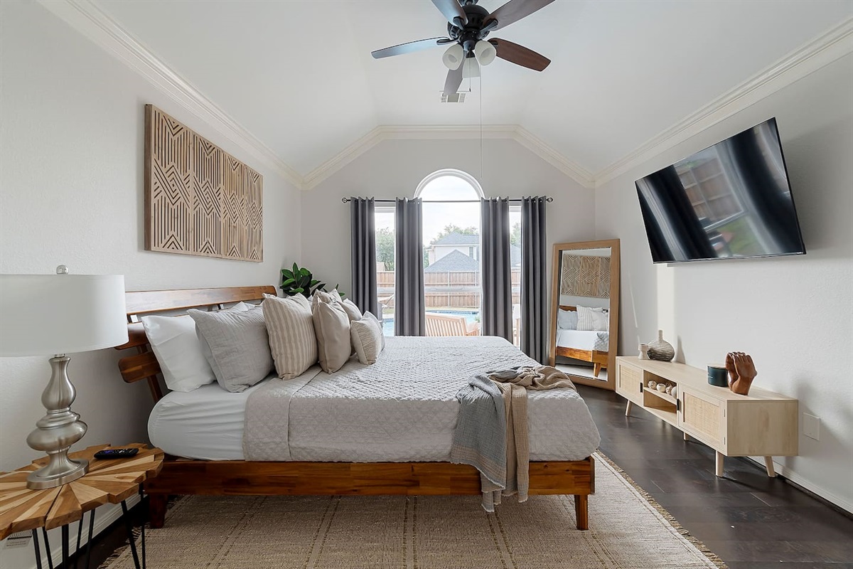 Rustic meets modern in this tranquil bedroom, with a grand bed, minimalist furnishings, and a sleek TV, all bathed in natural light.