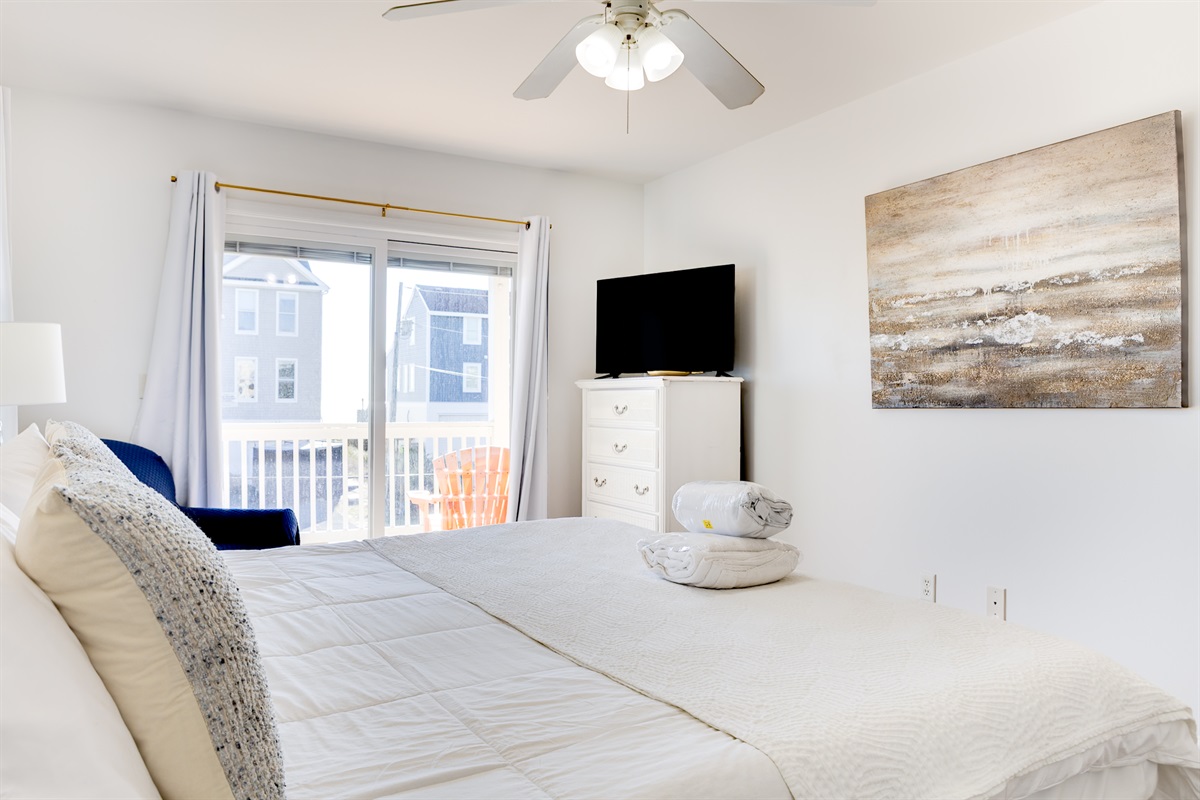 This airy master bedroom features a comfortable King bed with fresh linens, a ceiling fan, dresser with TV, and direct access to the balcony