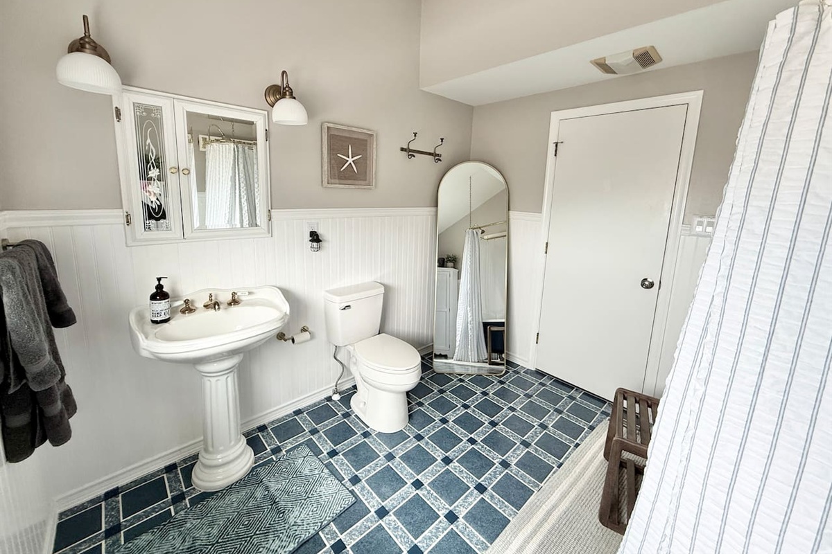 This spacious coastal-style bathroom features a white pedestal sink, blue and white checkered tile flooring, and nautical decor like a starfish and arched mirror.