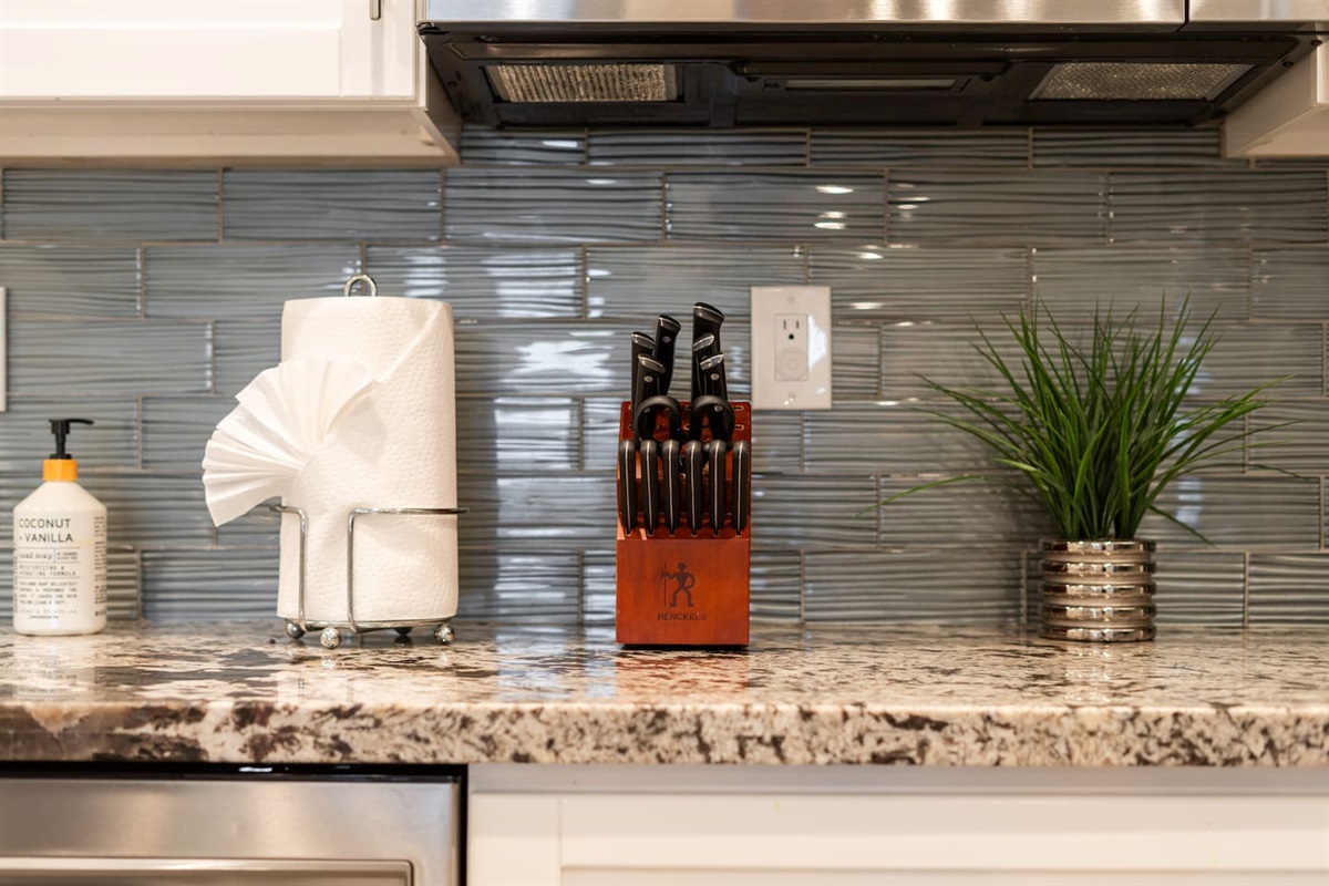 Close-up of a modern kitchen counter with granite surfaces, a glossy tile backsplash, and neatly arranged essentials, including a knife block and greenery.