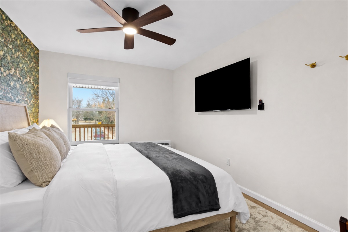 Bright, comfortable bedroom with a plush bed, smart TV, ceiling fan, and natural light—ideal for relaxing nights and easy mornings.