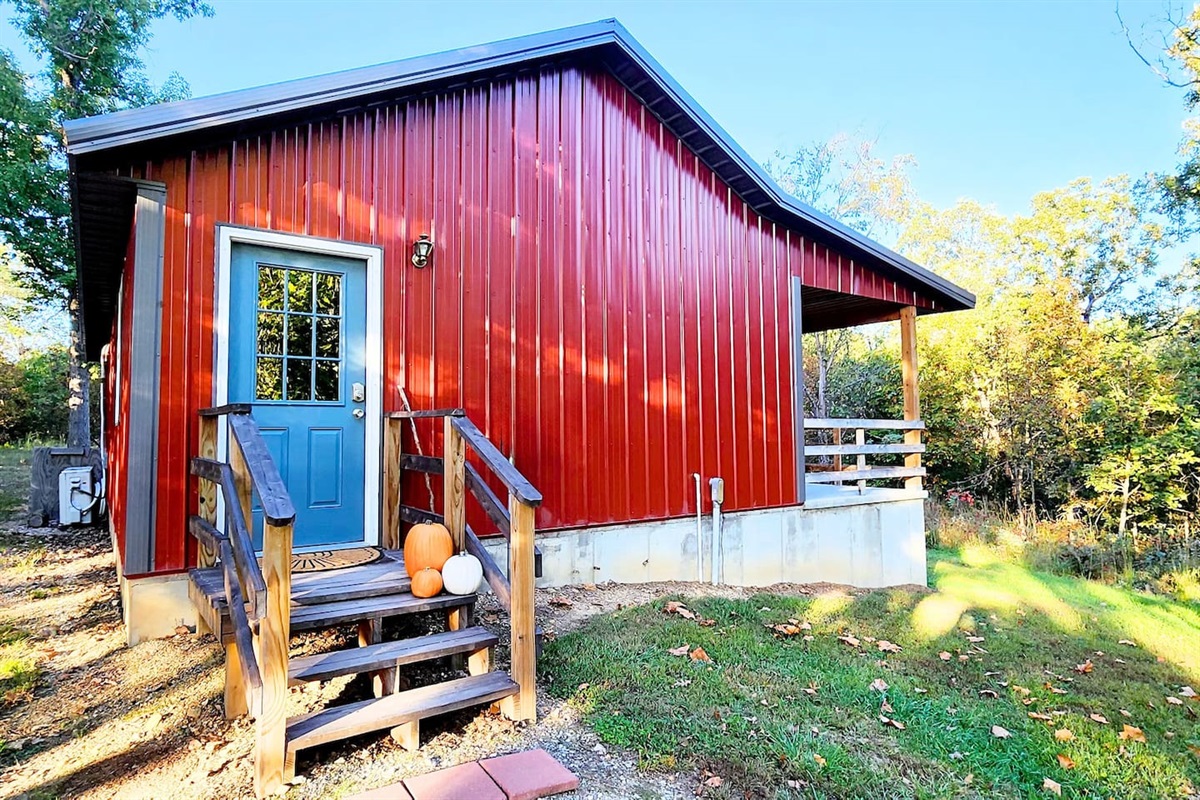 A vibrant red cabin glows in golden autumn light, with a blue front door and a few cheerful pumpkins welcoming you home. Surrounded by trees and nature, the cozy steps lead to a quiet porch—perfect for peaceful mornings or winding down at sunset