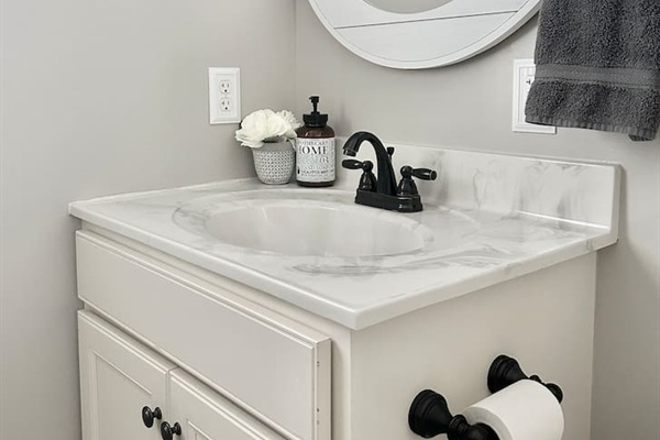 This bathroom vanity features a marbled white countertop with a recessed oval sink and a dark, two-handle faucet. The white cabinet base has dark knobs, and a matching dark toilet paper holder is mounted to its side.