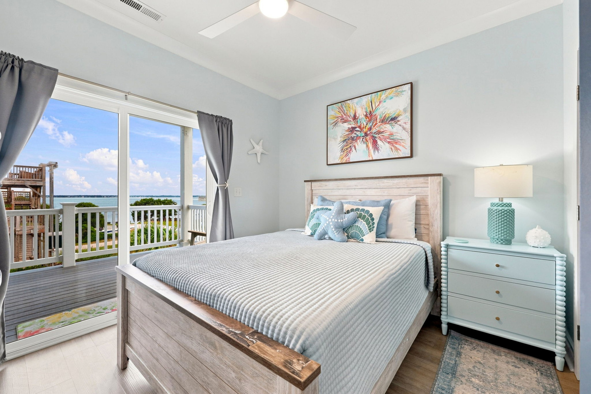 Bright second-floor bedroom featuring a comfortable queen bed, coastal-inspired decor, and beautiful water views from the private balcony