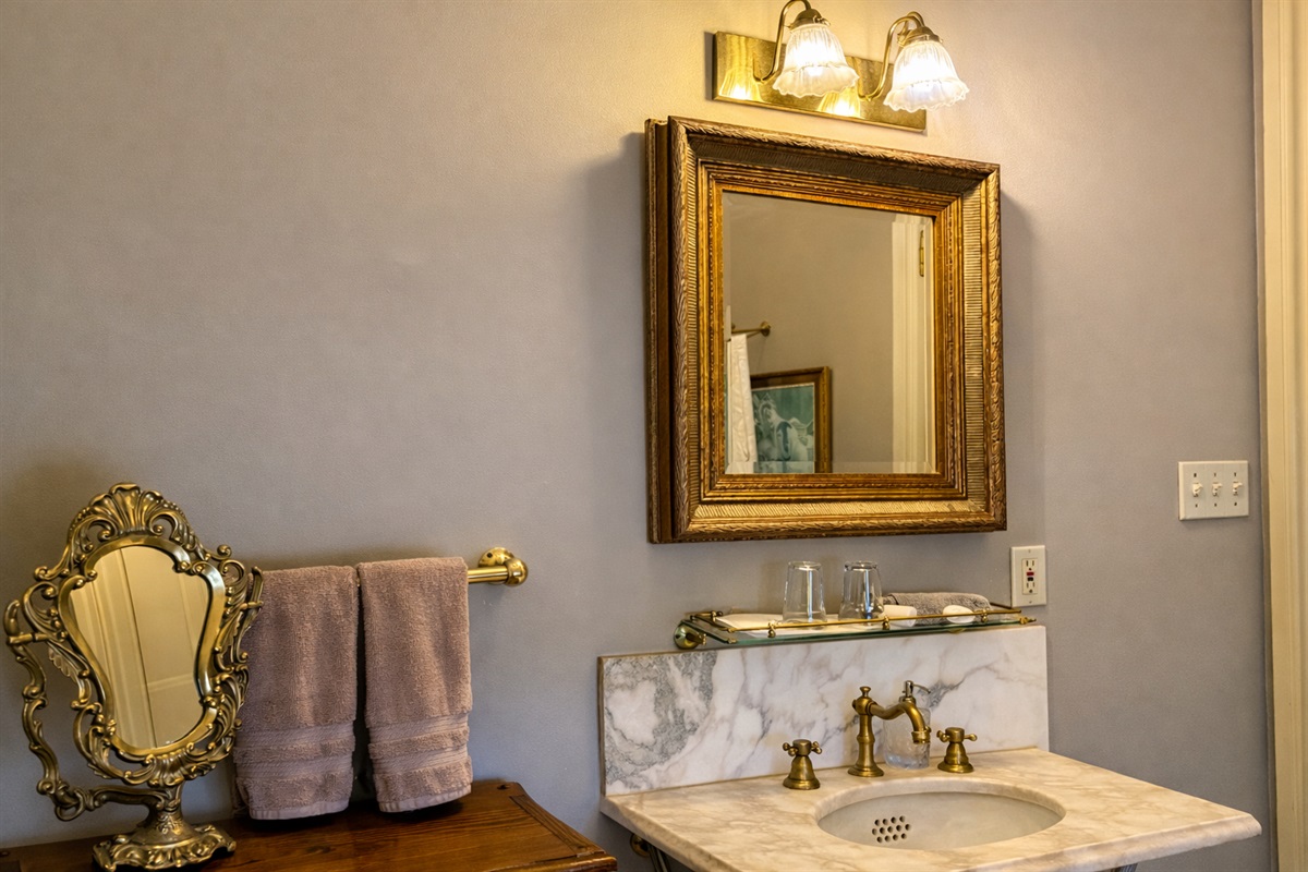 Bathroom vanity with marble sink, brass fixtures, framed mirror, and period-style lighting.