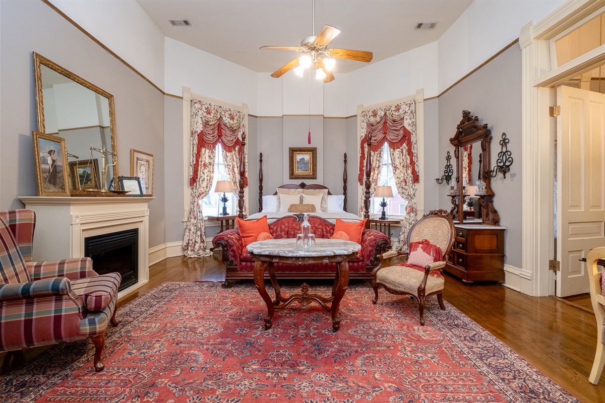 Victorian seating area with decorative fireplace, antique tables, and tall windows.