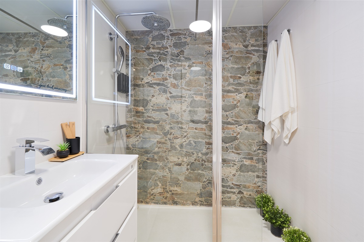 Modern bathroom with walk-in rain shower, stone accent wall, LED mirror, and fresh spa-like touches for your comfort.