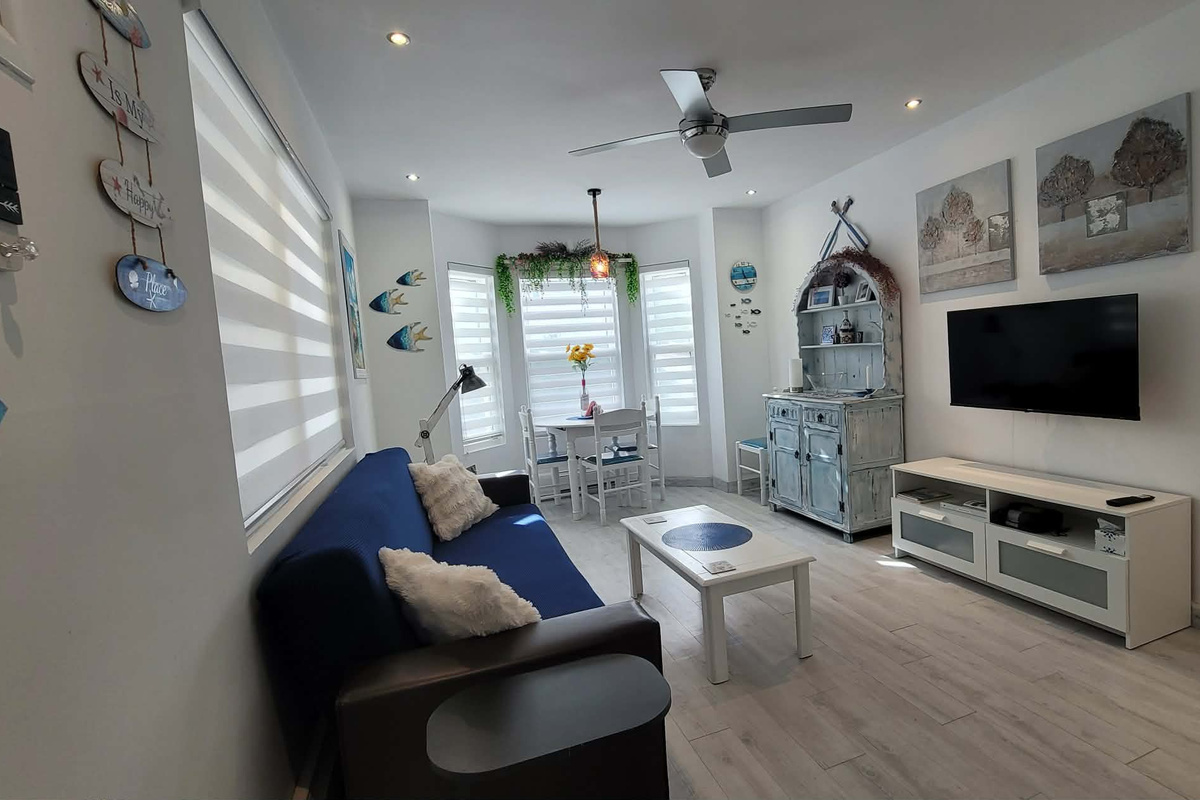 Living area with smart TV, ceiling fan and Air Conditioner, dining table for 4 guests, and futon sofa that folds out to a single bed for 1 adult or 2 small children.