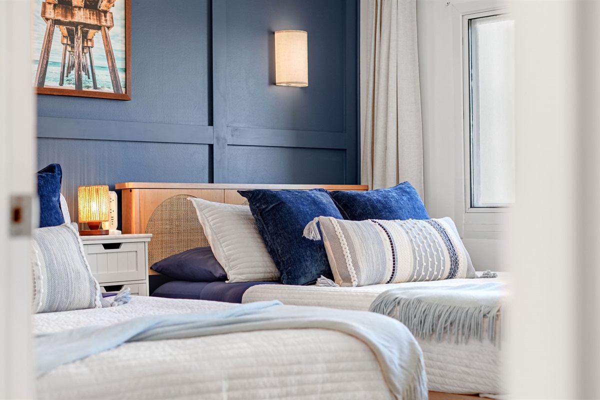 The bedroom features 2 queen beds, storage baskets for throw pillows and blankets, cordless sconces, blackout curtains, luxury bedding and stunning beach view from the panoramic window