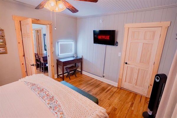 King bedroom featuring a wall-mounted smart TV, modern lighted vanity, and cozy bedding. Bright, functional setup for relaxing or getting ready.