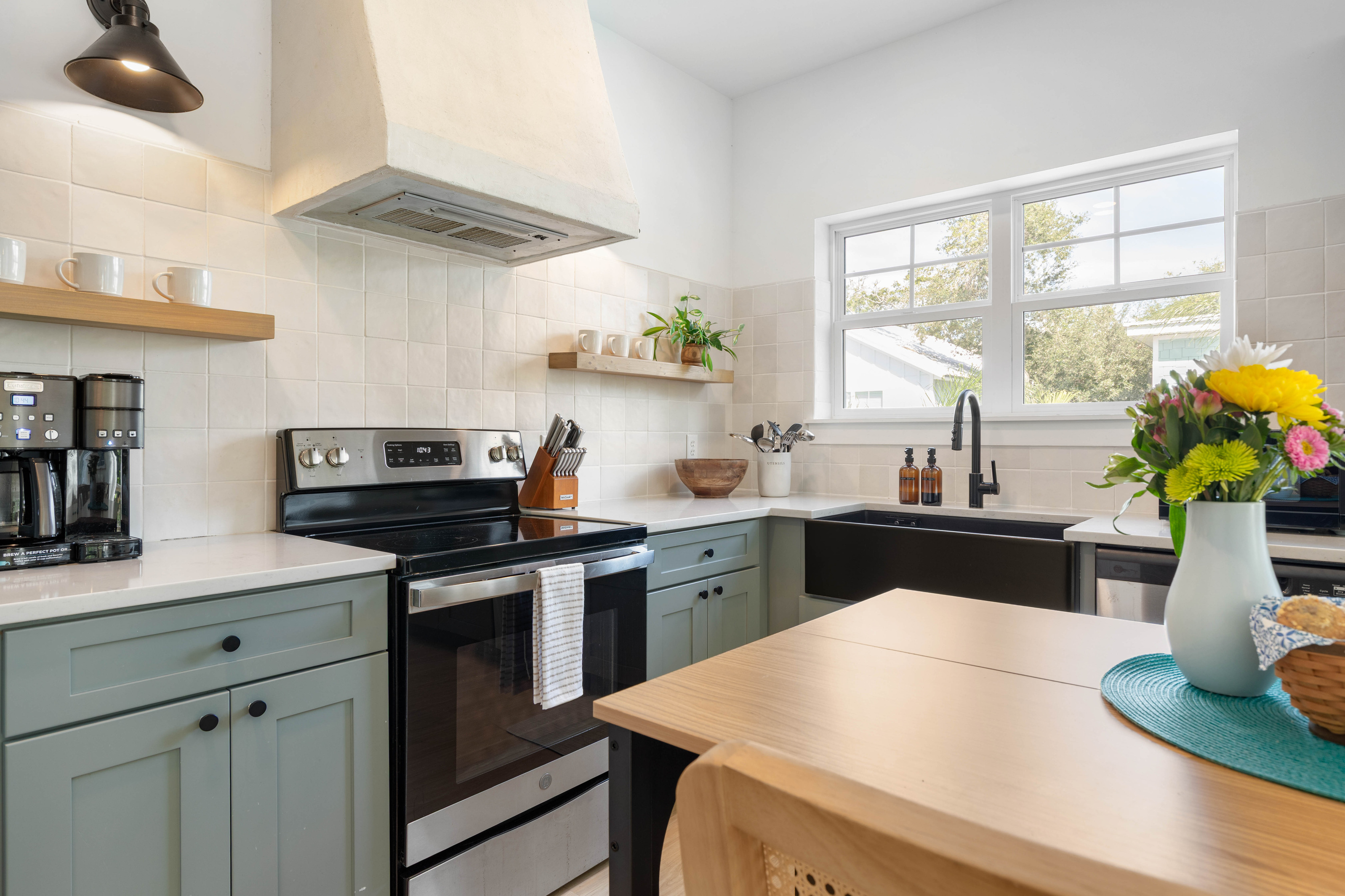 Two coffee makers on the counter, because there are two kinds of people on every family trip. The deep black sink takes corn husks and sandy buckets alike, and the window above it looks straight into the oaks.