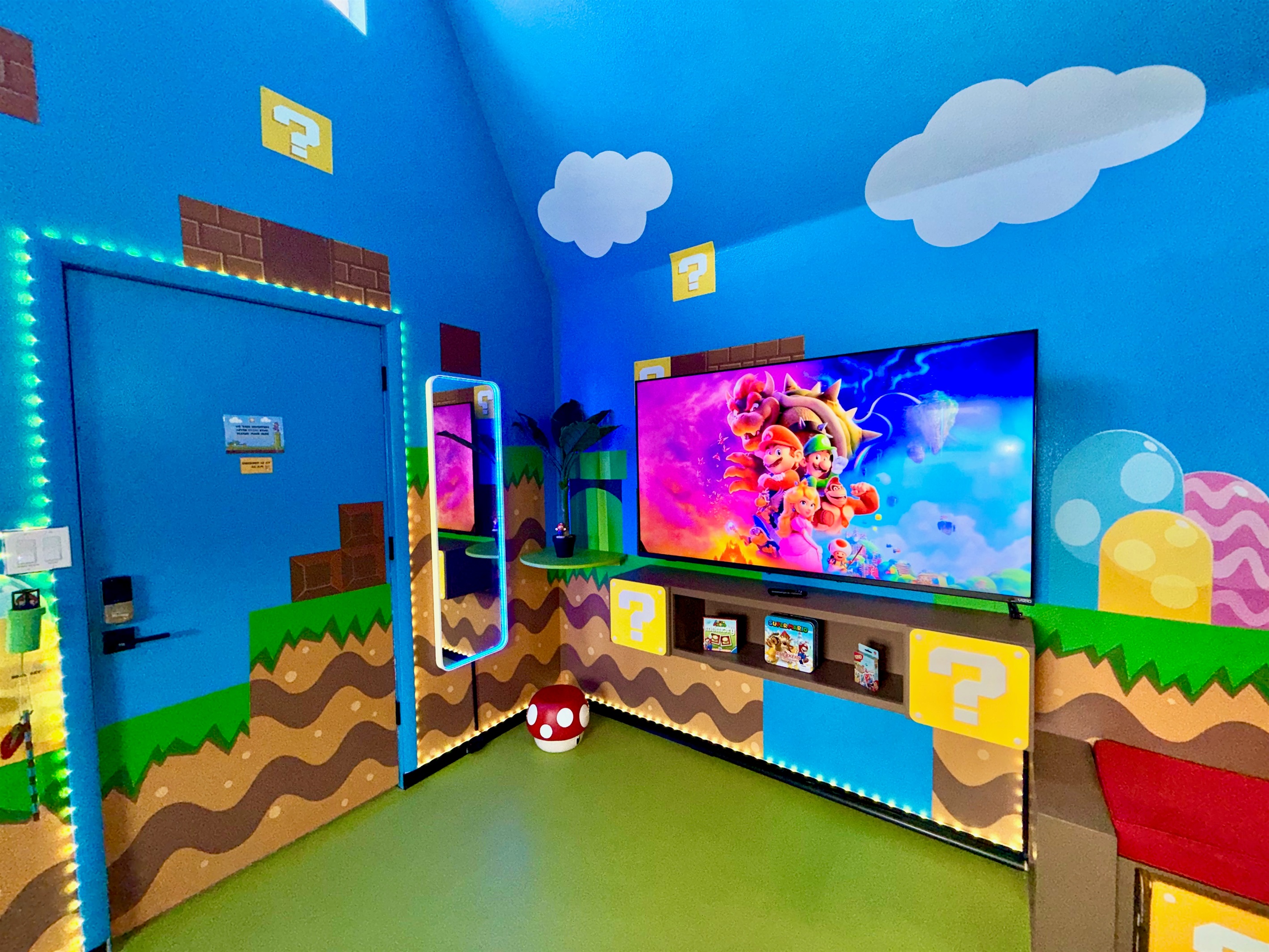 Vibrant blue walls with Super Mario murals, glowing question mark blocks, green pipes, and brick details surround the extra large 75" smart TV (+ Nintendo Switch system). The mirror lights up and plays music via Bluetooth, perfect for family fun!